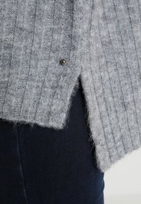 Grå stickad cardigan med ribbad textur, med en subtil glänsande knappdetalj på sidan och en något ojämn fåll.