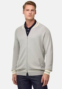 Cardigan zip grigio chiaro con colletto e polsini a coste, caratterizzato da righe crema, texture a maglia e tasche laterali. Indossato con una polo blu navy.
