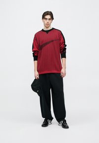 Piros-fekete csíkos pulóver V-nyakú kialakítással, laza fekete nadrággal és fekete sportcipővel párosítva. Egyik kézben fekete sapkát tart.