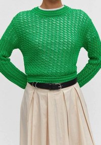 Grüner Strickpullover mit einem strukturierten, offenen Gewebe, kombiniert mit einem beigen Faltenrock und einem schwarzen Gürtel. Runder Ausschnitt und lange Ärmel.