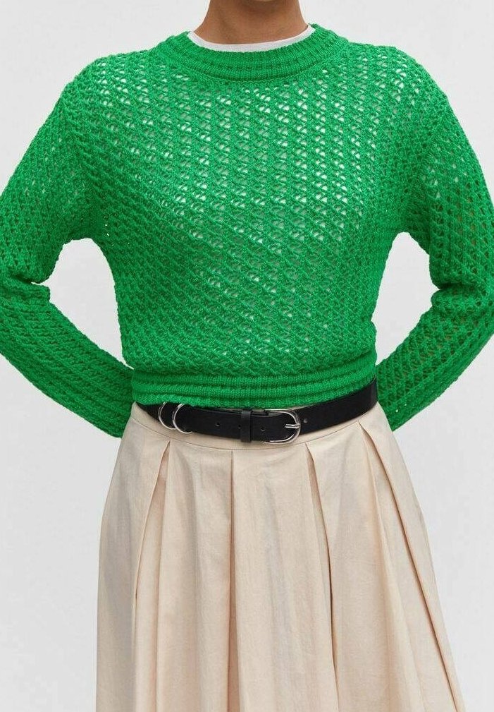 Grüner Strickpullover mit einem strukturierten, offenen Gewebe, kombiniert mit einem beigen Faltenrock und einem schwarzen Gürtel. Runder Ausschnitt und lange Ärmel.