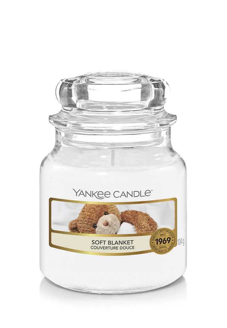 Yankee Candle con un tarro de vidrio, cera blanca y una etiqueta que presenta un oso de peluche marrón. El texto de la etiqueta dice "Manta Suave" con detalles dorados.