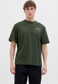Mørkegrøn bomulds t-shirt med korte ærmer, rund hals og et lille trykt logo på venstre bryst. Glat tekstur, afslappet pasform.