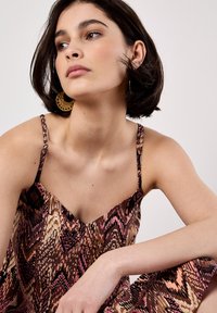 Primo piano di una giovane donna con capelli corti e scuri, che indossa un vestito a fantasia e grandi orecchini, mentre fissa pensierosa verso un lato.