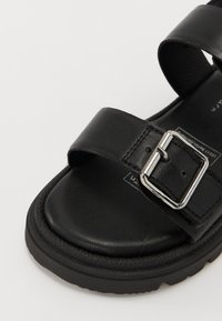 Sandal preto em couro com duas tiras largas e um fecho prateado. Sola grossa preta e textura suave com detalhes de costura visíveis.