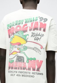 Λευκό μπλουζάκι φτιαγμένο από βαμβάκι που διαθέτει μια χρωματιστή γραφικά εικόνα ενός βάτραχου να παίζει κιθάρα, με κείμενο "FOREST HILLS '99 FROG JAM" και λεπτομέρειες εκδήλωσης παρακάτω.