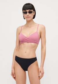 Rózsaszín bikini felsőrész keresztbe rakott dizájnnal és vékony pántokkal, amelyet egyszínű fekete bikini alsóval párosítanak. Simaság, minimalista stílus.