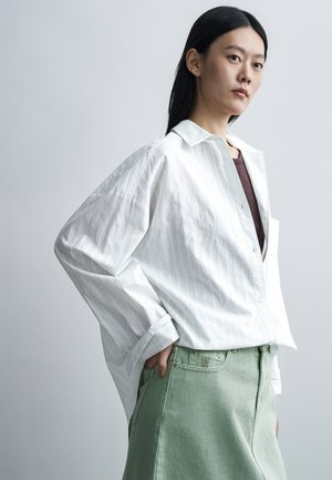Jeune femme aux longs cheveux noirs portant une chemise blanche ample boutonnée sur un haut foncé et une jupe vert clair, posant avec la main dans la poche.