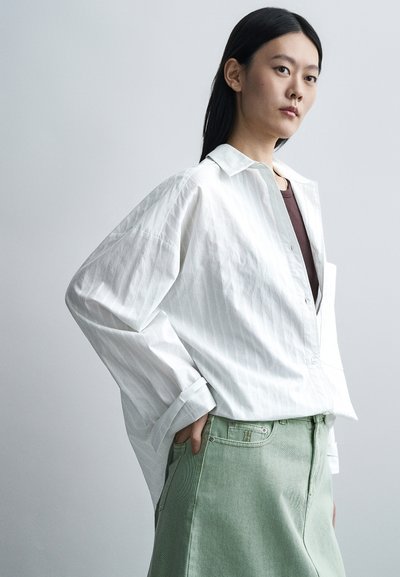 Jeune femme aux longs cheveux noirs portant une chemise blanche ample boutonnée sur un haut foncé et une jupe vert clair, posant avec la main dans la poche.