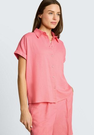 Vrouw met een loszittend, kort mouw roze overhemd met knopen en bijpassende roze short, staand met één hand in de zak.