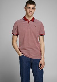 Rode polo met een effen textuur, contrasterende donkerrode kraag en blauwe en witte gestreepte accenten op de mouwen. Draagt met donkere spijkerbroek.