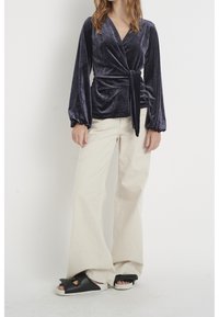 Tumma violetti samettinen wrap-top, jossa on pitkät, puffattuja hihoja, sidottuna vyötärölle. Yhdistetty vaaleanbeigeihin leveälahkeisiin housuihin ja mustiin liukusandaaleihin.