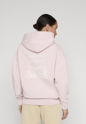 Mujer con cabello oscuro recogido en un moño, vestida con una sudadera con capucha de color rosa claro con una ilustración de una tienda de flores y pantalones beige, de espaldas contra un fondo liso.