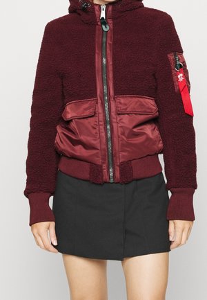 Veste bourgogne avec une texture pelucheuse, un devant en satin, deux grandes poches avant et une fermeture éclair. Elle comprend une capuche et des poignets en tricot.