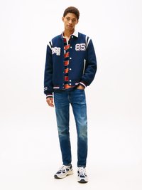 Chaqueta de varsity navy con rayas blancas, que presenta parches y el número 85. Lleva puesta sobre una camiseta de rayas rojas y navy, combinada con jeans azules y zapatillas.