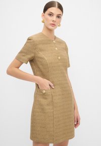 Kortärmad, figurnära klänning i ett strukturerat, beige tyg. Utrustad med guldfärgade knappar och två framskjutna fickor. Minimalistisk design med rund halsringning.
