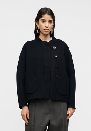 Granatowa marynarka wełniana z zaokrąglonym kołnierzem, wyposażona w duże naszywane kieszenie i pięć brązowych guzików z przodu. Miękka w dotyku i luźnego kroju.