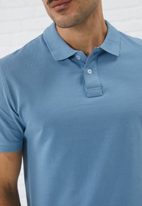 Polo bleu clair en tissu de coton texturé. Présente une patte de boutonnage à trois boutons et un design de col classique.