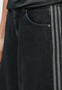 Close-up of black denim pants with triple gray side stripes and a blue-trimmed front pocket, worn with a black top.