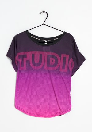 Lyhythihainen urheilupaita violetin ja pinkin liukuvärjäyksellä, jossa iso ääriviivateksti "STUDIO" rinnan kohdalla, mustalla henkarilla ripustettuna.