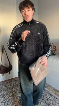 Veste noire en tissu pelucheux avec fermeture éclair, rayures blanches sur les manches, sac gris à la main, et pantalon ample foncé. Fond neutre avec décoration.