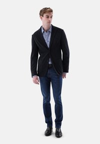 Schwarzer Cord-Blazer über einem gemusterten blauen Hemd, kombiniert mit dunkelblauen Jeans und schwarzen Schuhen. Verfügt über einen Zwei-Knopf-Verschluss und Vordertaschen.