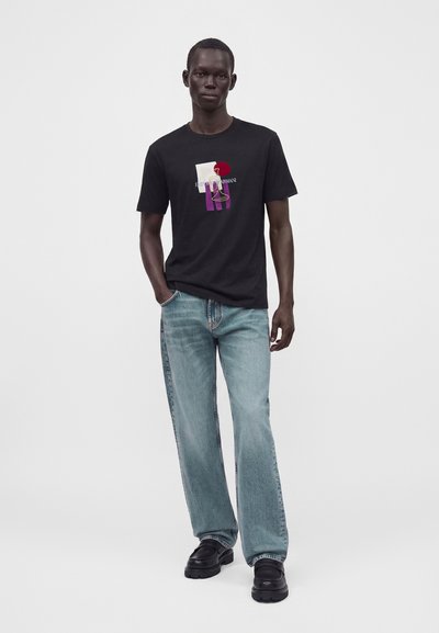 Άνδρας με μαύρο γραφικό t-shirt, ανοιχτό μπλε τζιν και μαύρα παπούτσια, που στέκεται με το ένα χέρι στην τσέπη μπροστά σε απλό φόντο.
