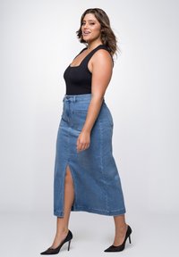 Denimrok met een zijsplit, voorzien van een hoge taille en voorzakken. G gedragen met een zwarte aansluitende body en zwarte puntige hakken.