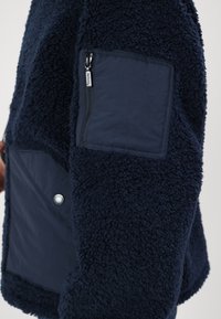 Marineblaue Fleecejacke mit strukturiertem Außenmaterial, ausgestattet mit einer Reißverschlusstasche und Druckknopfdetails auf der Vorderseite.