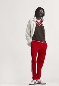 Červené teplákové nohavice s bielymi bočnými pruhmi, hnedý sveter s červenými akcentmi, biela bunda a biele tenisky s červenými pruhmi. Strieborné slnečné okuliare.