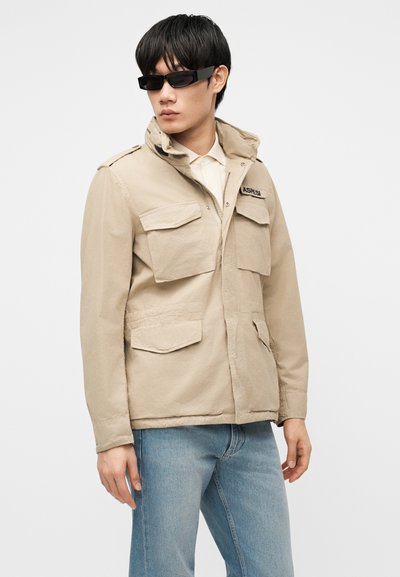 Homme portant une veste utilitaire beige avec plusieurs poches avant, jean bleu clair, chemise blanche et lunettes de soleil rectangulaires noires, debout devant un fond blanc.