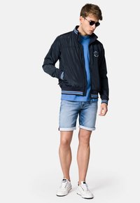 Granatowa kurtka bombowca z białymi i niebieskimi paskami na mankietach, w zestawieniu z jasnoniebieską koszulką i dżinsowymi szortami. Białe sneakersy dopełniają look.