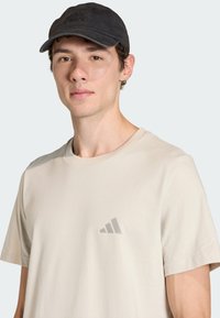 Beżowa bawełniana koszulka z delikatnym szarym logo Adidas na piersi, w połączeniu z czarną czapką z logo Terrex o fakturowanym wykończeniu.