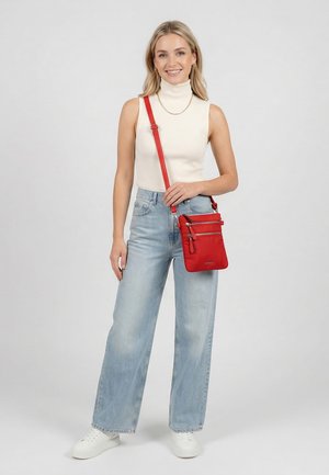 Junge Frau in ärmellosem weißem Rollkragenpullover und hellblauen Jeans, die eine rote Umhängetasche mit mehreren Reißverschlusstaschen hält.