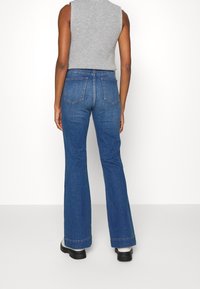 Modré rozšířené džíny s vysokým pasem, designem pěti kapes a jemným prošíváním. Nosí se s šedým bezrukávovým topem a černou obuví.