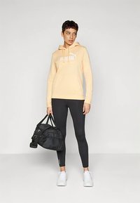 Gaiši persiku krāsas sporta džemperis ar baltu logotipu, melnas legingi, baltas sporta kurpes un melna sporta soma. Modelis stāv uz neitrālas krāsas fona.