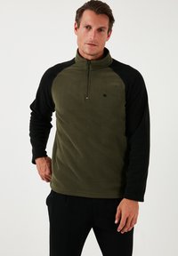 Olive zöld színű fleece pulóver fekete raglánujjakkal; félig cipzáros gallérral és kis logóval a mellrészen. Puha tapintású, laza szabású.