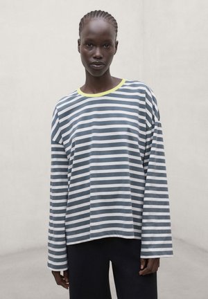 Persoon met gevlochten haar die een losse grijs-wit gestreepte longsleeve met een gele kraag draagt, staande tegen een effen lichte achtergrond.