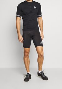 Černé cyklistické tričko s krátkým rukávem a zipem, se sektorováním bílé značky a pruhovanými akcenty. Spárováno s úzkými černými cyklistickými šortkami.