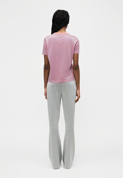 Γυναίκα με μακριά πλεξούδα, φορούν ροζ μπλούζα με κοντά μανίκια και ψηλόμεσο ανοιχτό γκρι παντελόνι με φαρδιά ποδάρια, στέκεται με την πλάτη προς τα εμπρός.