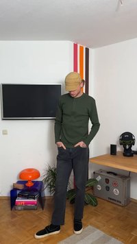 Camisa henley de mangas largas verde-escura com placket de botões, combinada com jeans pretos e sapatos slip-on pretos. Boné bege e acessórios minimalistas.