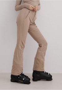 Beige skibukser med slim fit, med glidelåslommer og kontrastpaneler. Kombinert med svarte skistøvler som viser merkevare og utstyr.