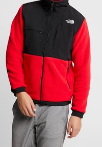 Giacca in pile con zip rossa e nera, colletto alto, tasche frontali e polsini elastici. Presenta colori a contrasto e una superficie tessuta.