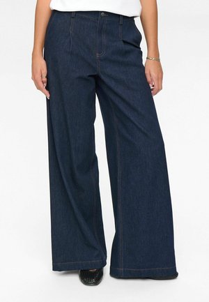 Woman wearing dark blue wide-leg denim jeans with visible orange stitching and black woven shoes, standing against a white background.