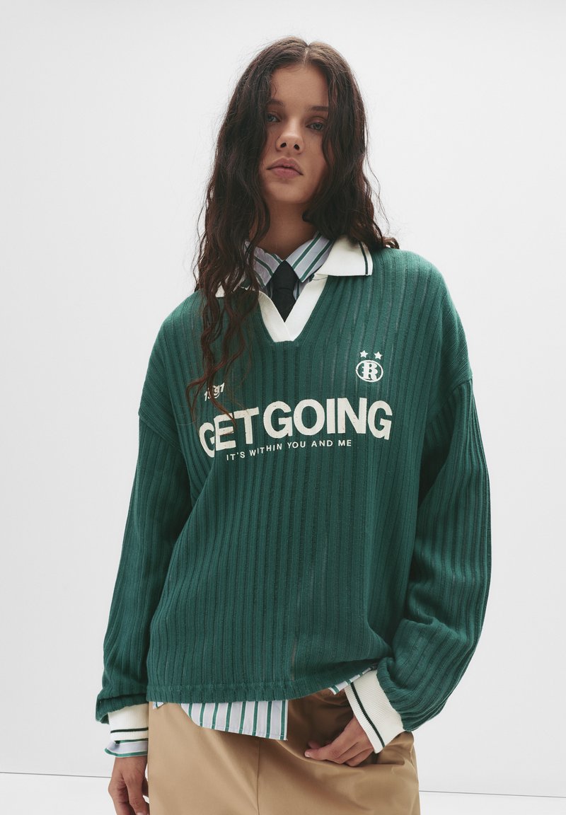 Zelená rebrovaná voľná mikina s výstrihom do V, biely nápis "GET GOING" a logo s hviezdami. Nosená cez košelu s golierom a prúžkami.