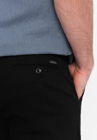 Pantalon noir en tissu texturé, avec une poche au dos, une fermeture à bouton et un petit accent logo en métal sur la taille.