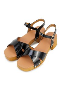 Zwarte lakleren sandalen met kruisbanden, houten blokhakken en een tan zool. Inclusief decoratieve gouden hardware accenten.