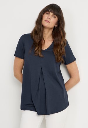 Marineblå t-shirt med korte ærmer og V-hals samt plisseret frontdetalje, lavet af blødt stof, kombineret med hvide bukser. Glat tekstur, afslappet pasform.