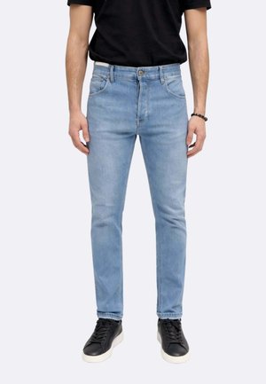 Uomo che indossa jeans aderenti di colore azzurro chiaro, T-shirt nera, sneakers nere con suole bianche, in piedi contro uno sfondo chiaro e semplice.