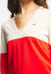 Svetr s véčkovým výstřihem a barevným blokovým designem, krémové a červené barvy, pletená textura, s malým zeleným logem na levé hrudi.